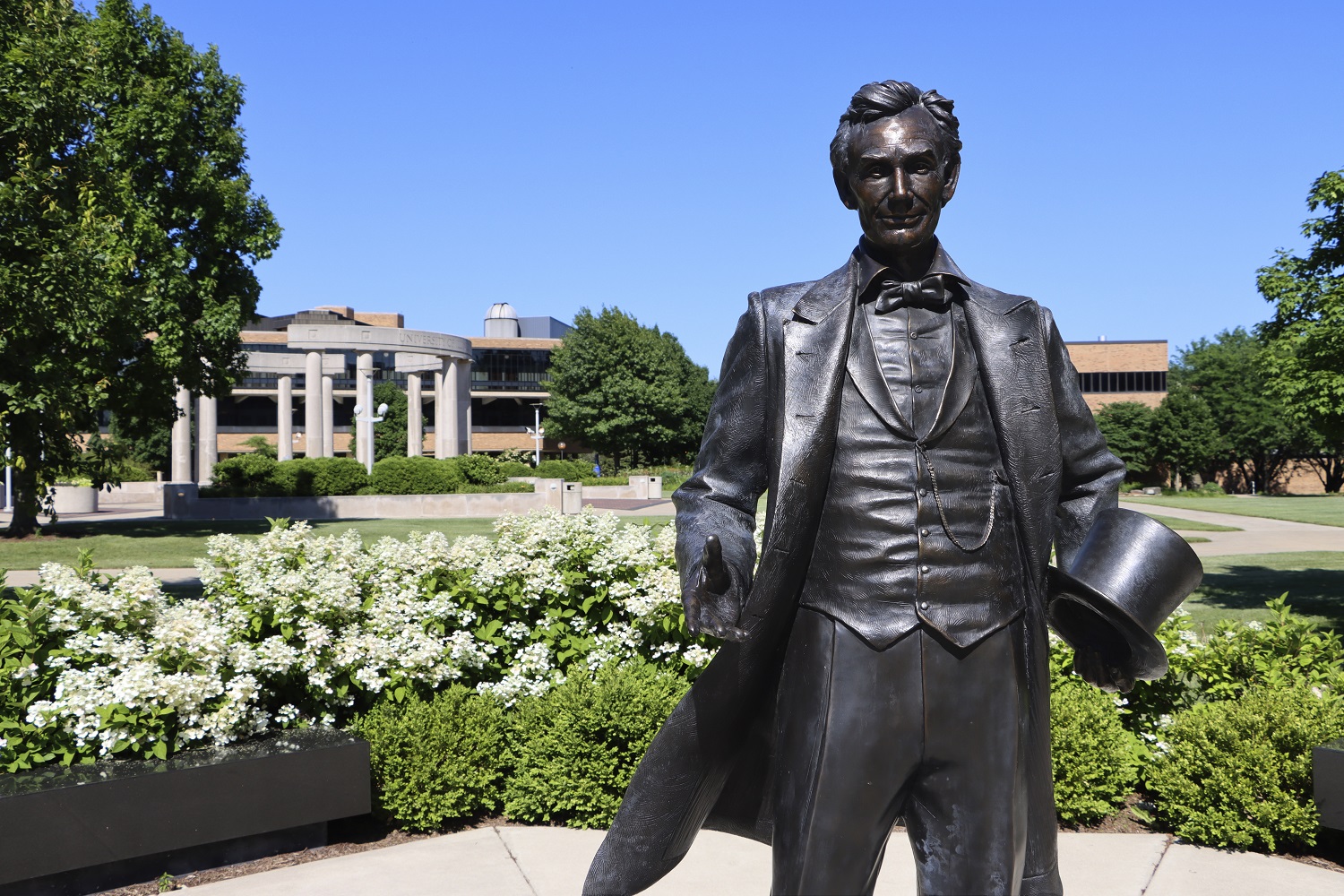 Photo of the UIS Young Lincoln Statue surrounded by flowering bushes with the UIS Colonnade in the background, taken by Blake Woods on July 1st, 2022.